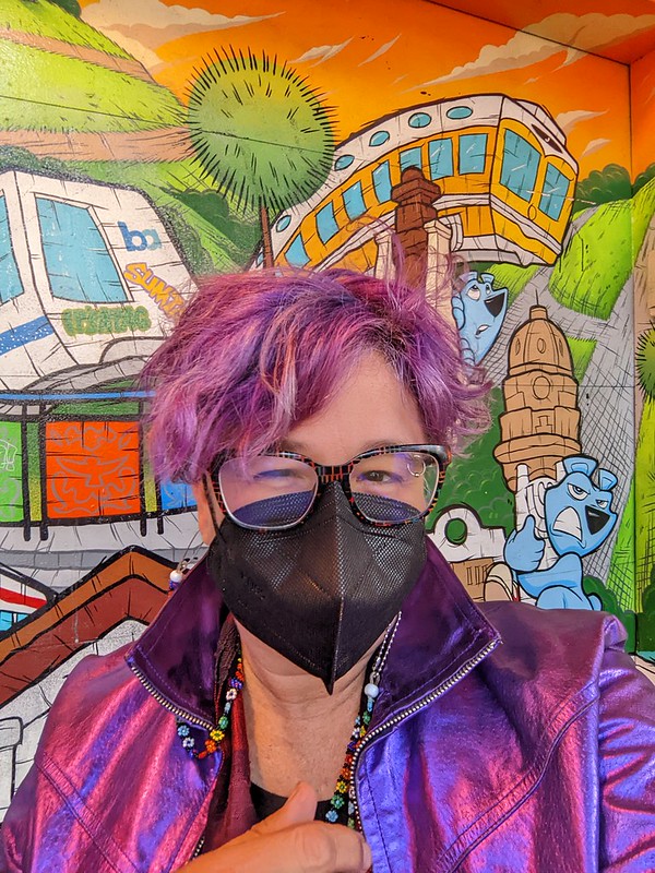 A purple haired white person with fabulous rainbow glasses